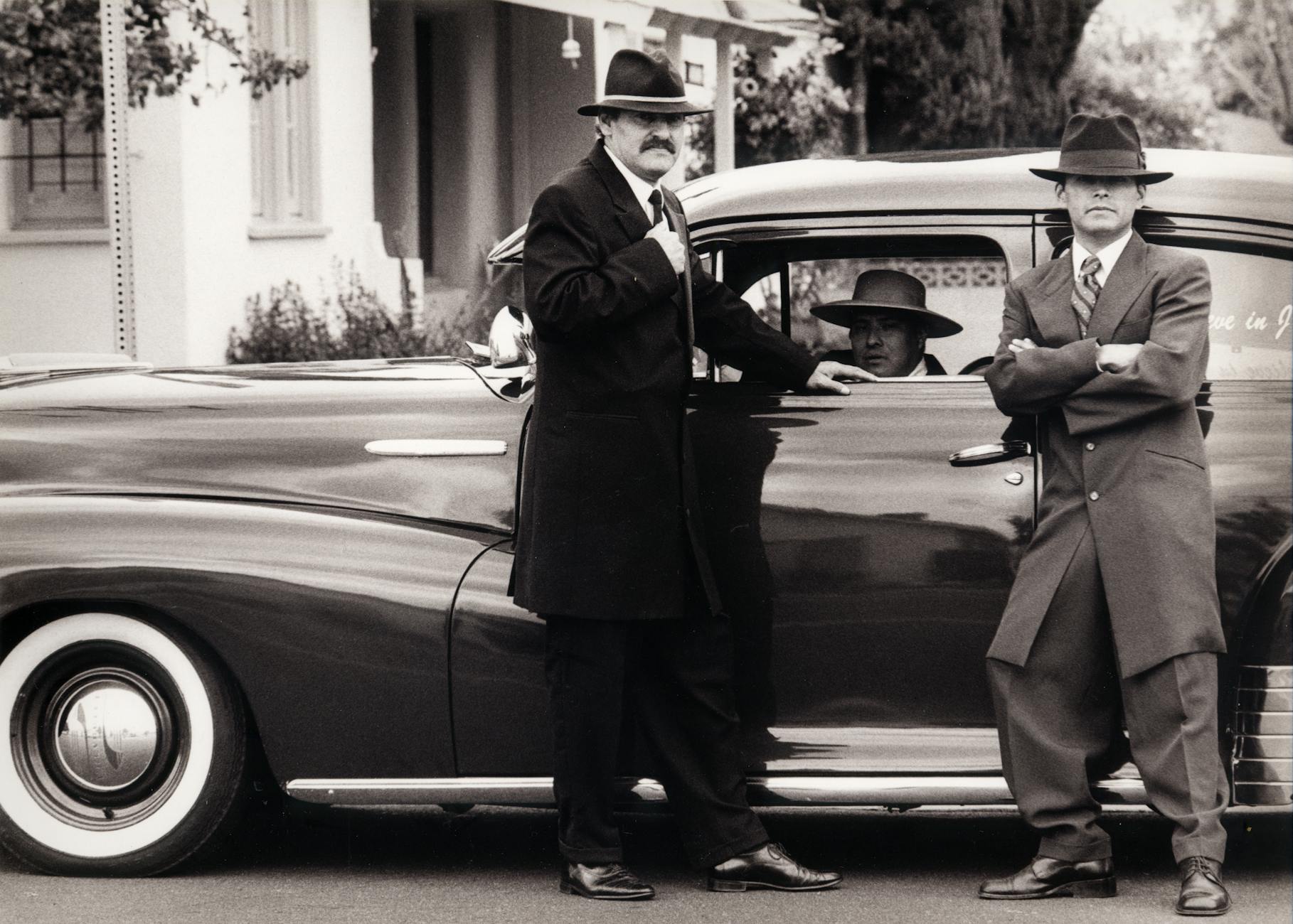 men wearing coats and fedoras standing in front of a car
